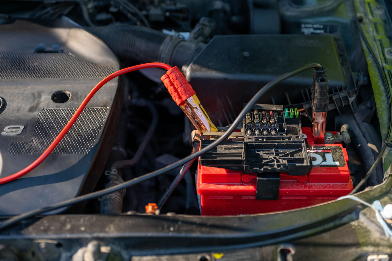 Car battery in an engine bay with red and black jumper cables clamped to its terminals