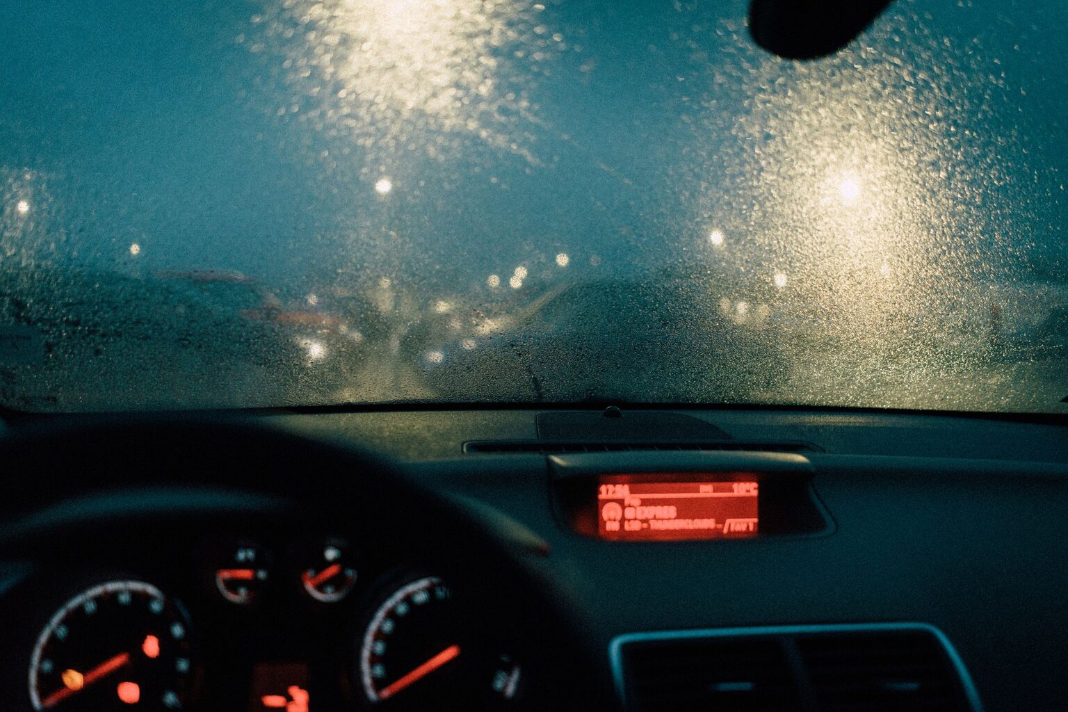 A rain-covered windshield at night with city lights blurred beyond it and the instrument panel lit below