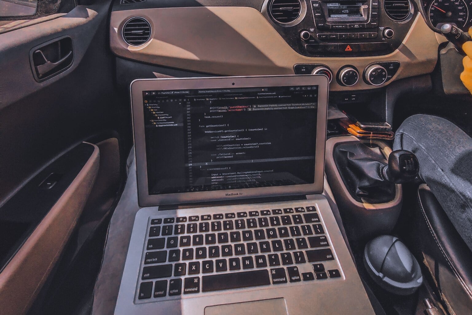 An open laptop on the passenger seat of a parked car, with the dashboard and gear lever alongside