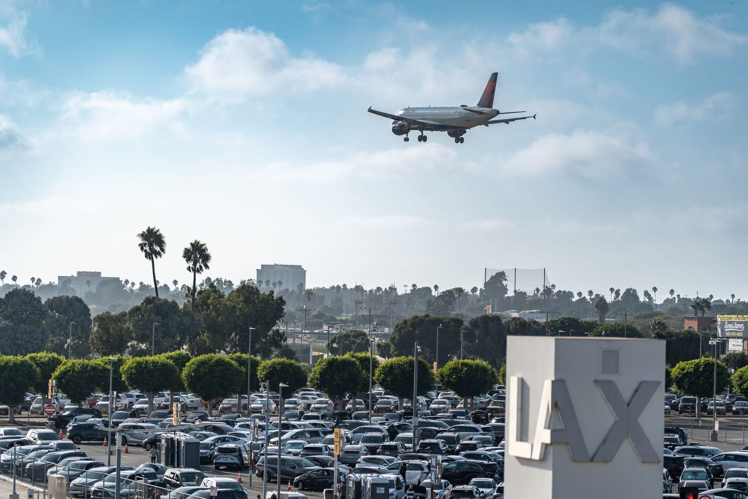 Plane landing over a large airport parking lot beside an LAX sign